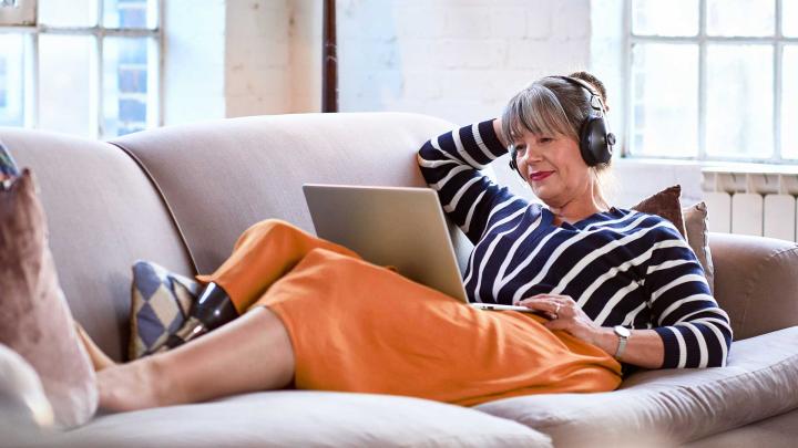 A woman wearing headphones sits on a couch and uses a laptop