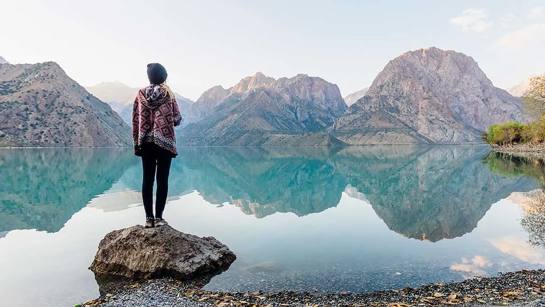 A person stands on a rock in the water and looks at the surrounding mountains